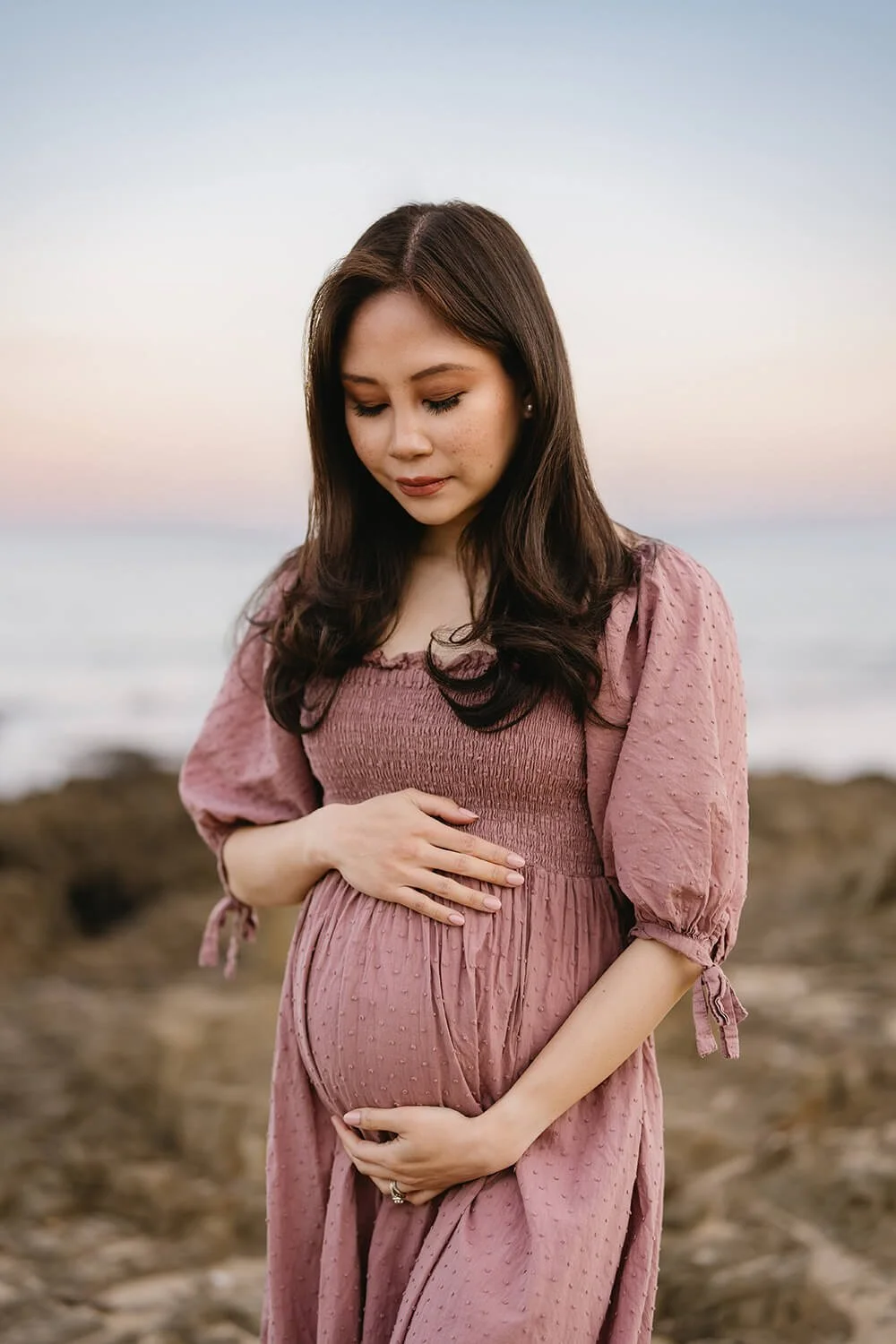 A pregnant woman in a pink dress with puffed sleeves and tie details on the sleeves stands on a rocky beach at sunset, gently holding her belly with one hand and resting her other hand on top of it, looking down with a serene expression.