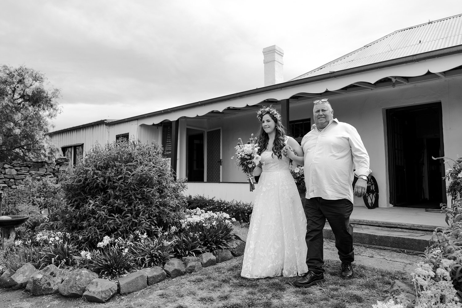 A bride in a white wedding dress with a floral crown holding a bouquet, walking arm-in-arm with a man in a white shirt and dark pants outside a house with a garden.