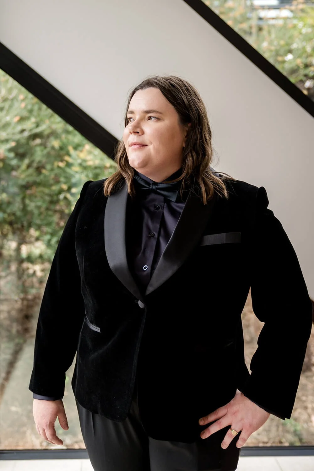 Person with shoulder-length brown hair, dressed in a black tuxedo with satin lapels, standing by a large window with greenery outside, posing confidently.