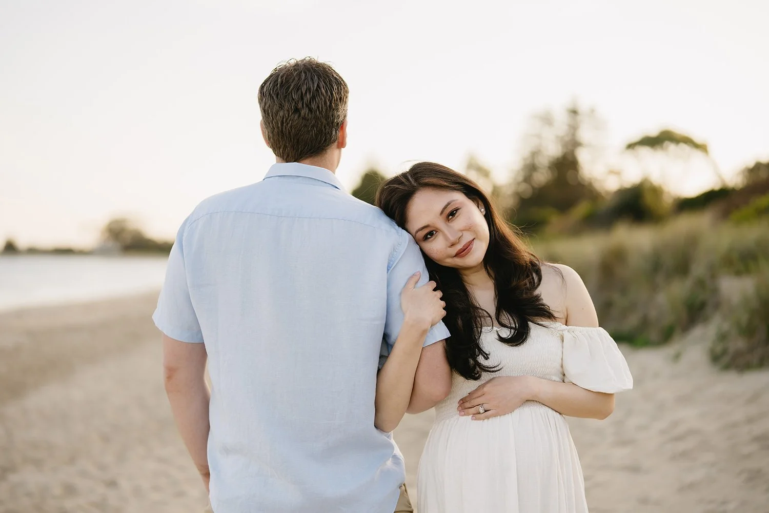 A woman leaning her head on a man's shoulder at the beach during sunset.