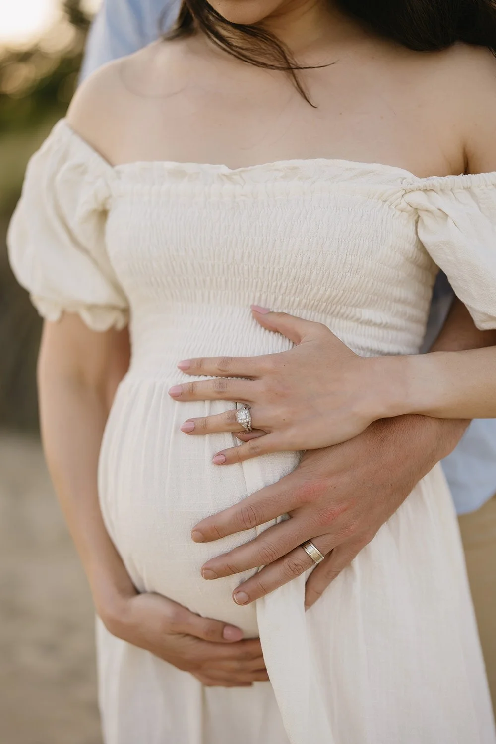 Close-up of a pregnant woman in a cream-colored off-the-shoulder dress with her hands resting on her belly, wearing wedding rings.
