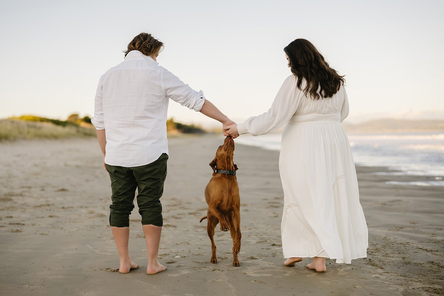 A couple holding hands with a dog on a beach during sunset, the woman is wearing a white dress and the man is in a white shirt and dark pants.