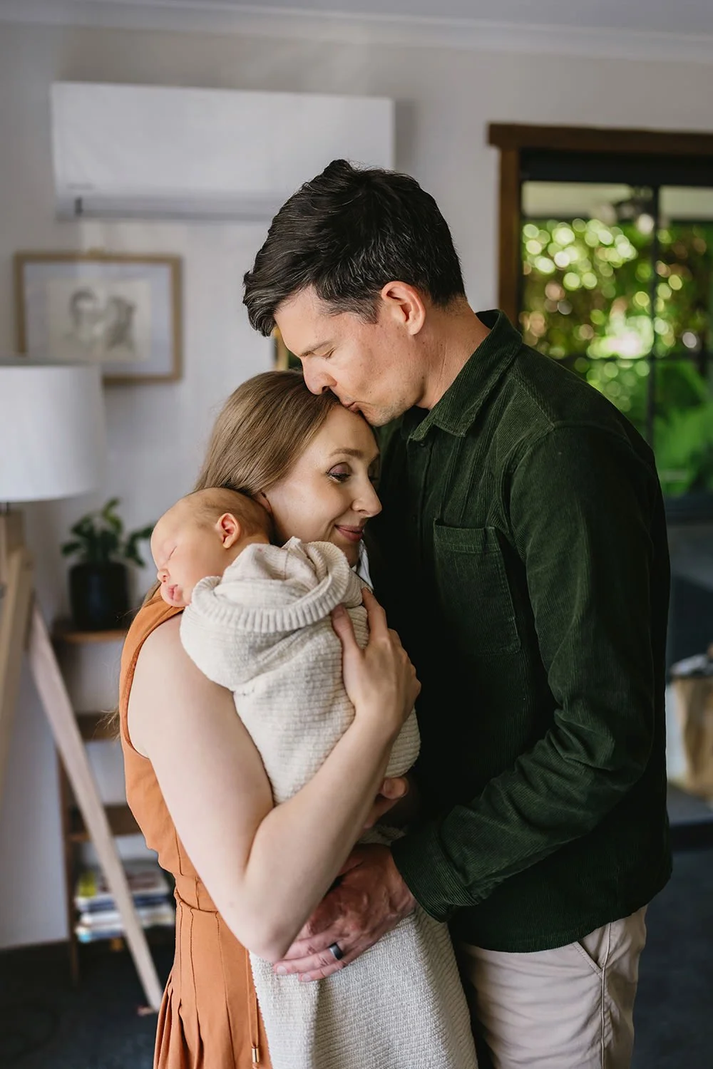 A family embracing at home, featuring a woman holding a newborn baby, a man kissing her forehead, with indoor decor and a window showing greenery in the background.
