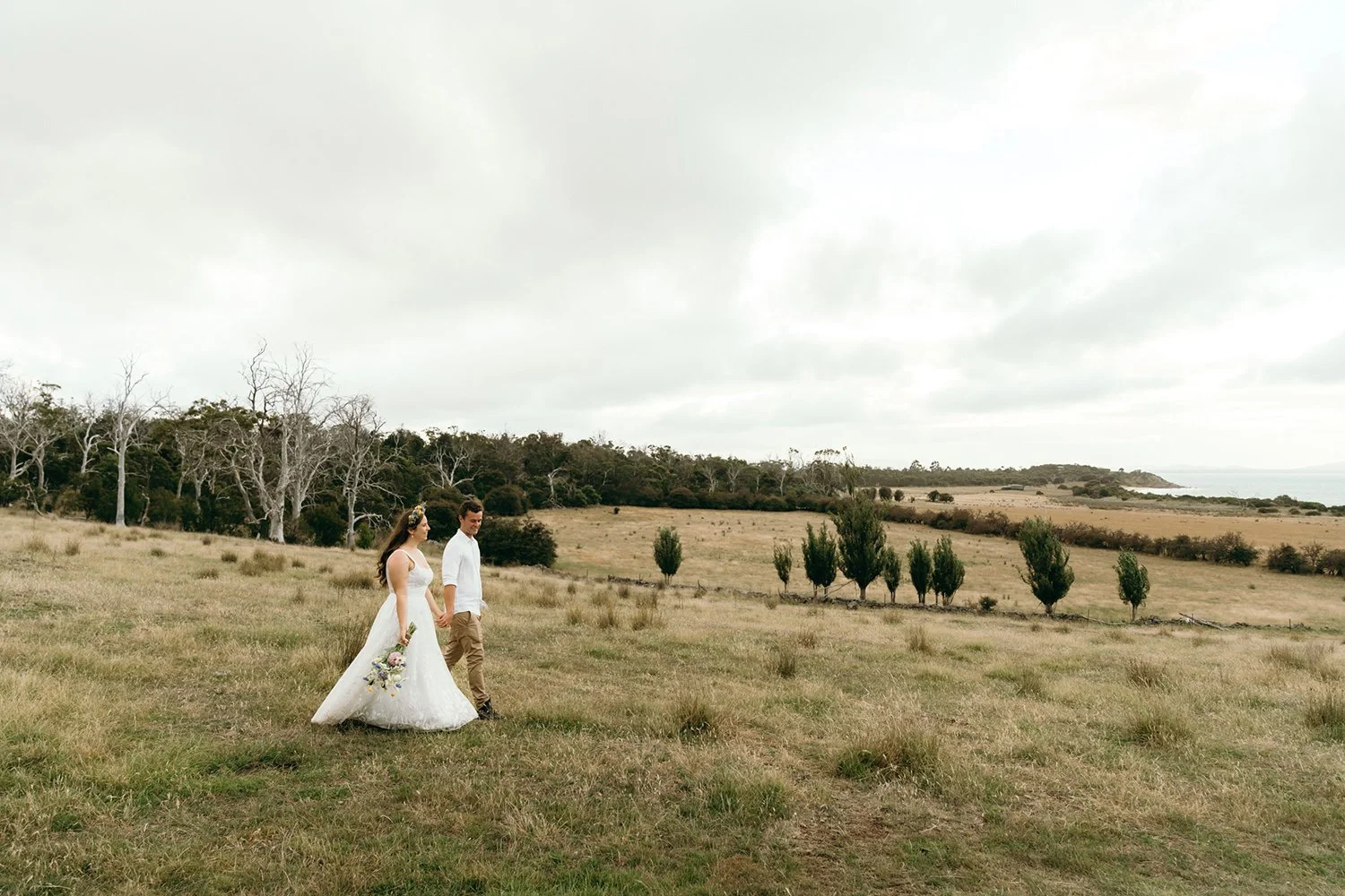 A bride and groom holding hands and walking through a grassy field on a cloudy day with scattered trees in the background.