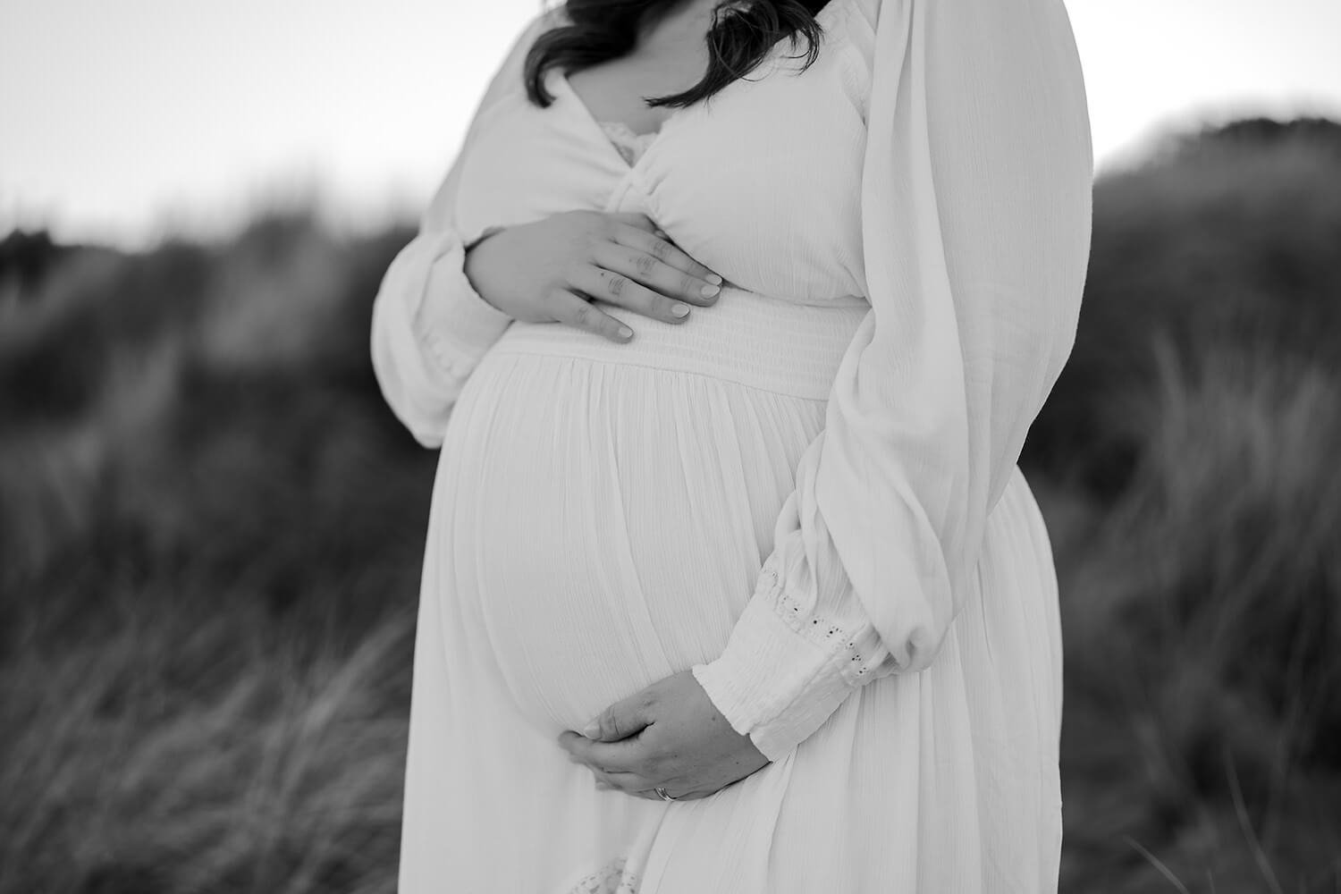 A pregnant woman dressed in white holds her belly with one hand and touches her chest with the other outdoors.