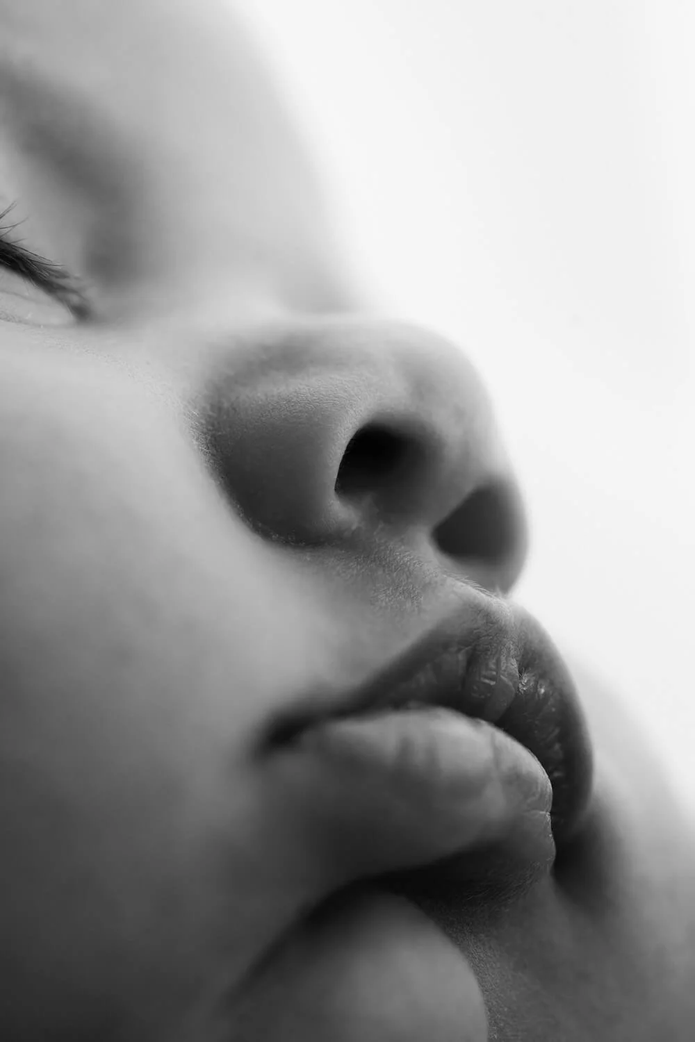 Close-up black and white photograph of a baby's face, focusing on the nose and lips, with part of one eye visible and the rest softly blurred.