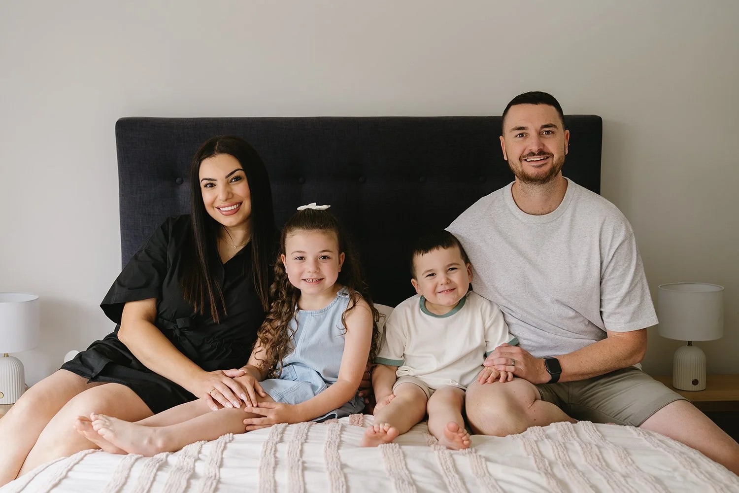A family of five sitting on a bed in a bedroom, smiling at the camera. The mother is on the left in a black dress, the father is on the right in a gray t-shirt, with two children, a girl and a boy, in between them. The girl has long curly hair and is