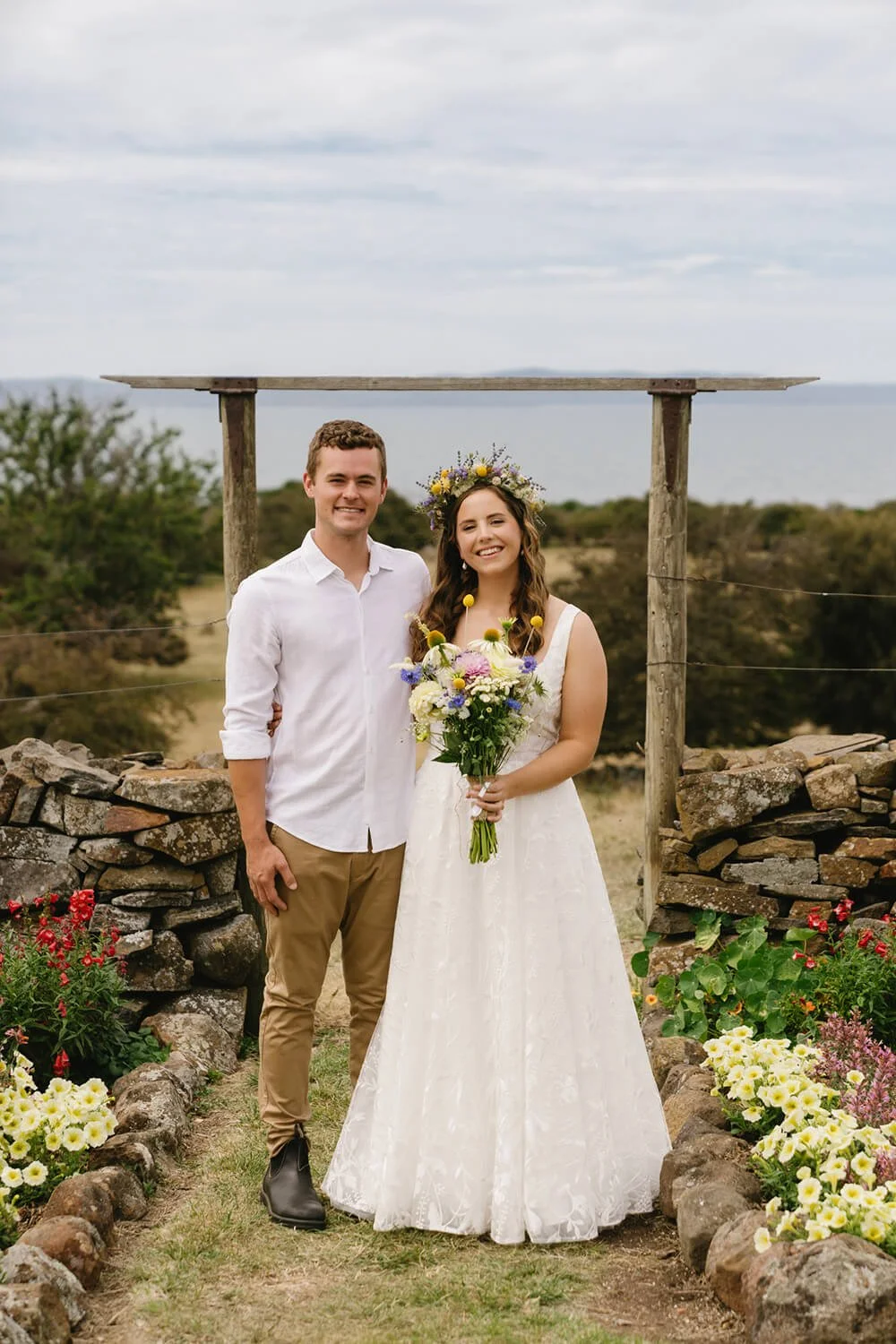 A couple celebrating their wedding outdoors with a stone and wood arch, surrounded by flowers and greenery, against a backdrop of sky and distant water.