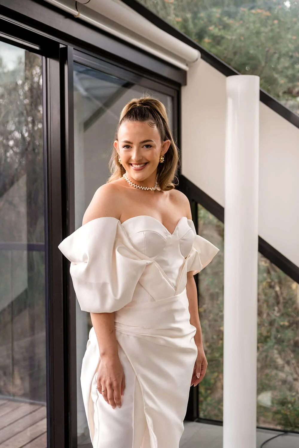 A woman in an off-shoulder white dress with puffed sleeves and jewelry, smiling inside a modern building with large glass windows and a view of trees outside.