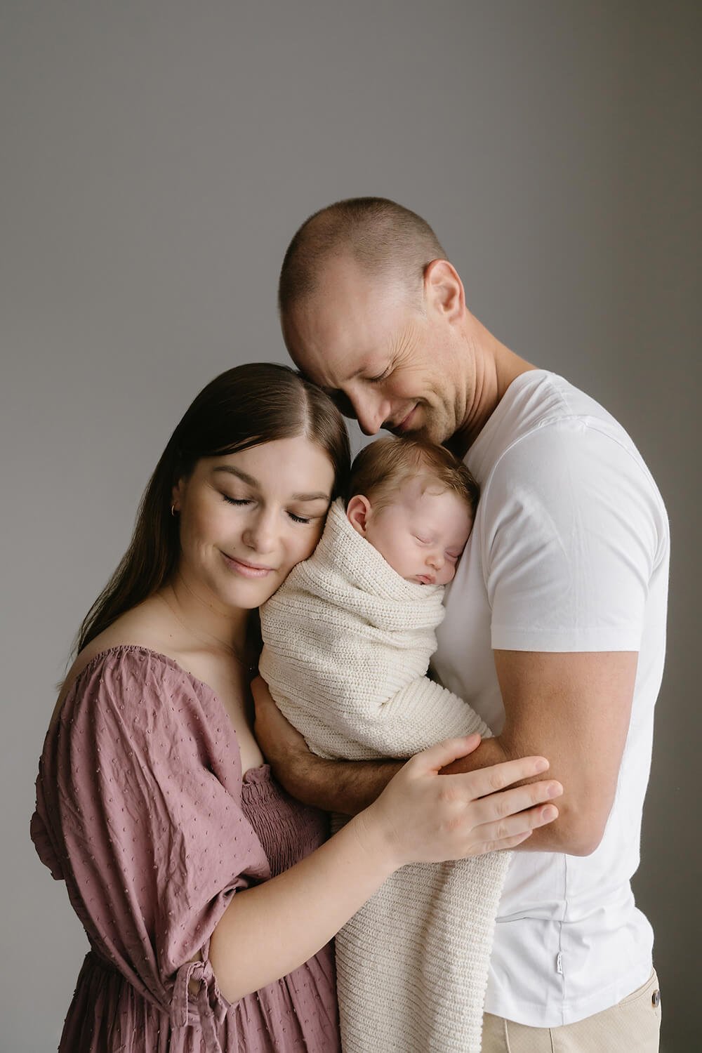 A family of three, a mother, father, and a newborn baby, are embracing each other and smiling gently with closed eyes, in a neutral-toned studio setting.