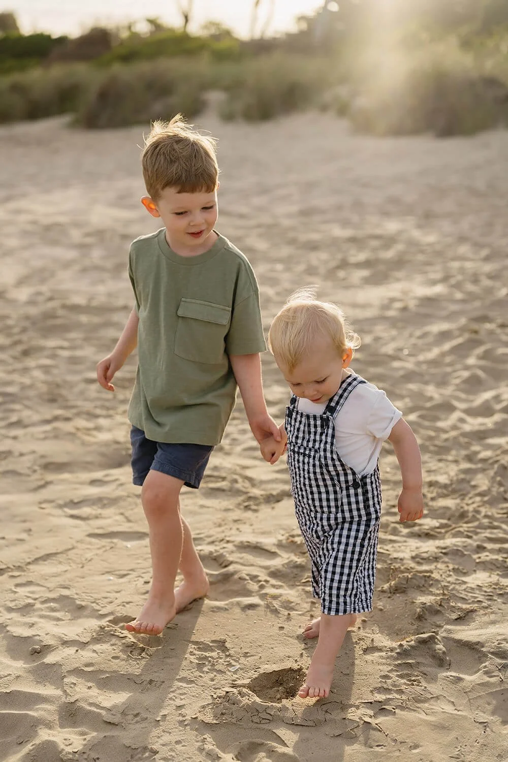 Two young boys walking hand in hand on a sandy beach during sunset.