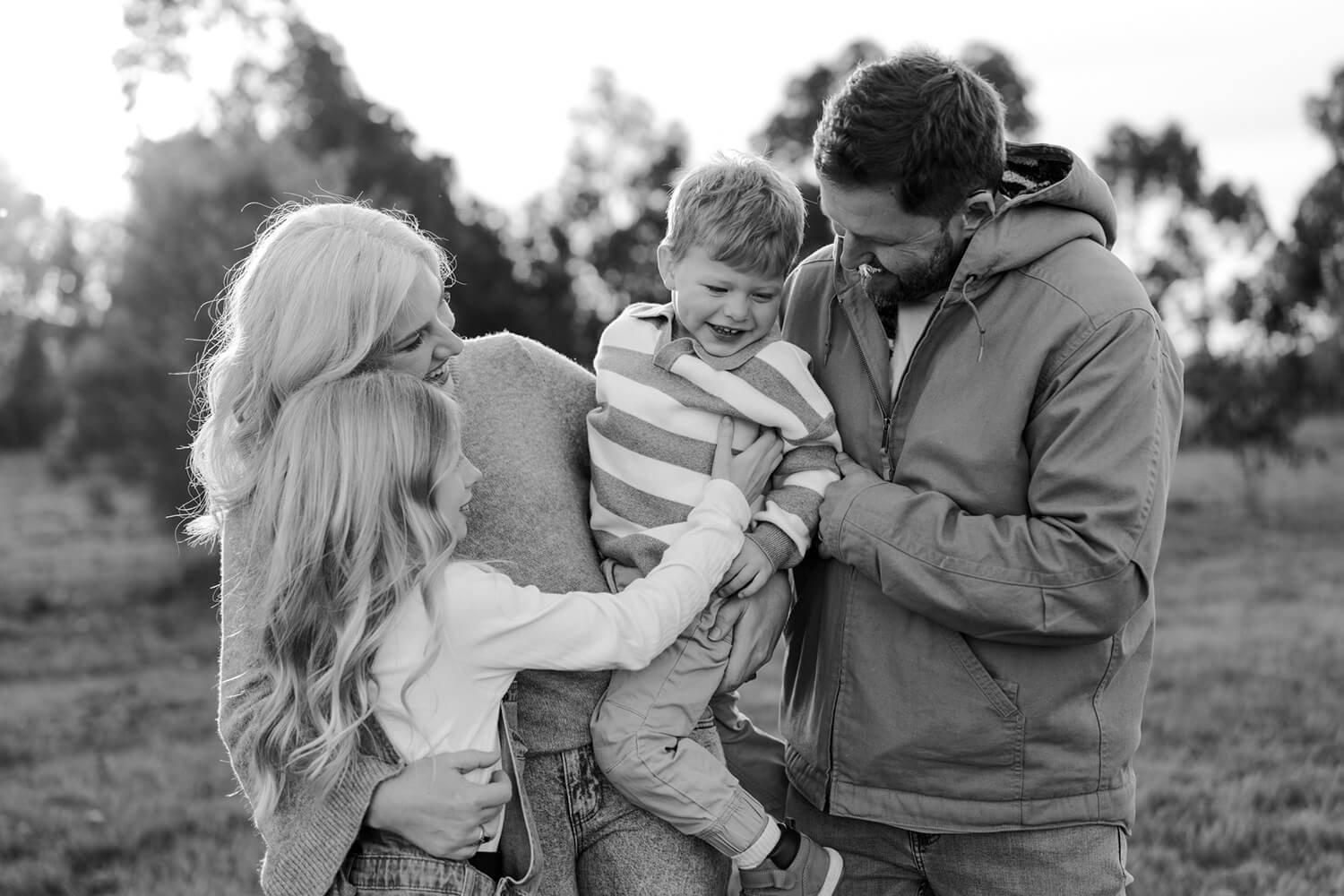 A family of four outdoors, with two women and two children. The children, a boy in a striped sweater and a girl with long curly hair, are being held and smiled at by the adults. The family appears happy and enjoying each other's company.
