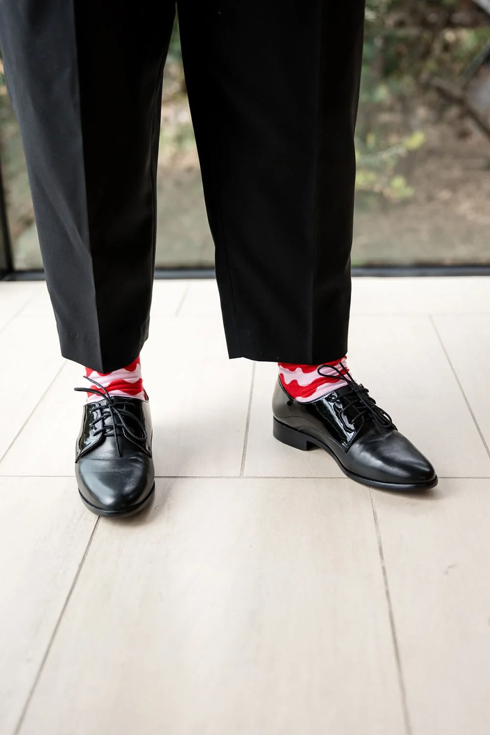 Person wearing black leather shoes, black pants, and red and white patterned socks, standing on light-colored tiled floor.