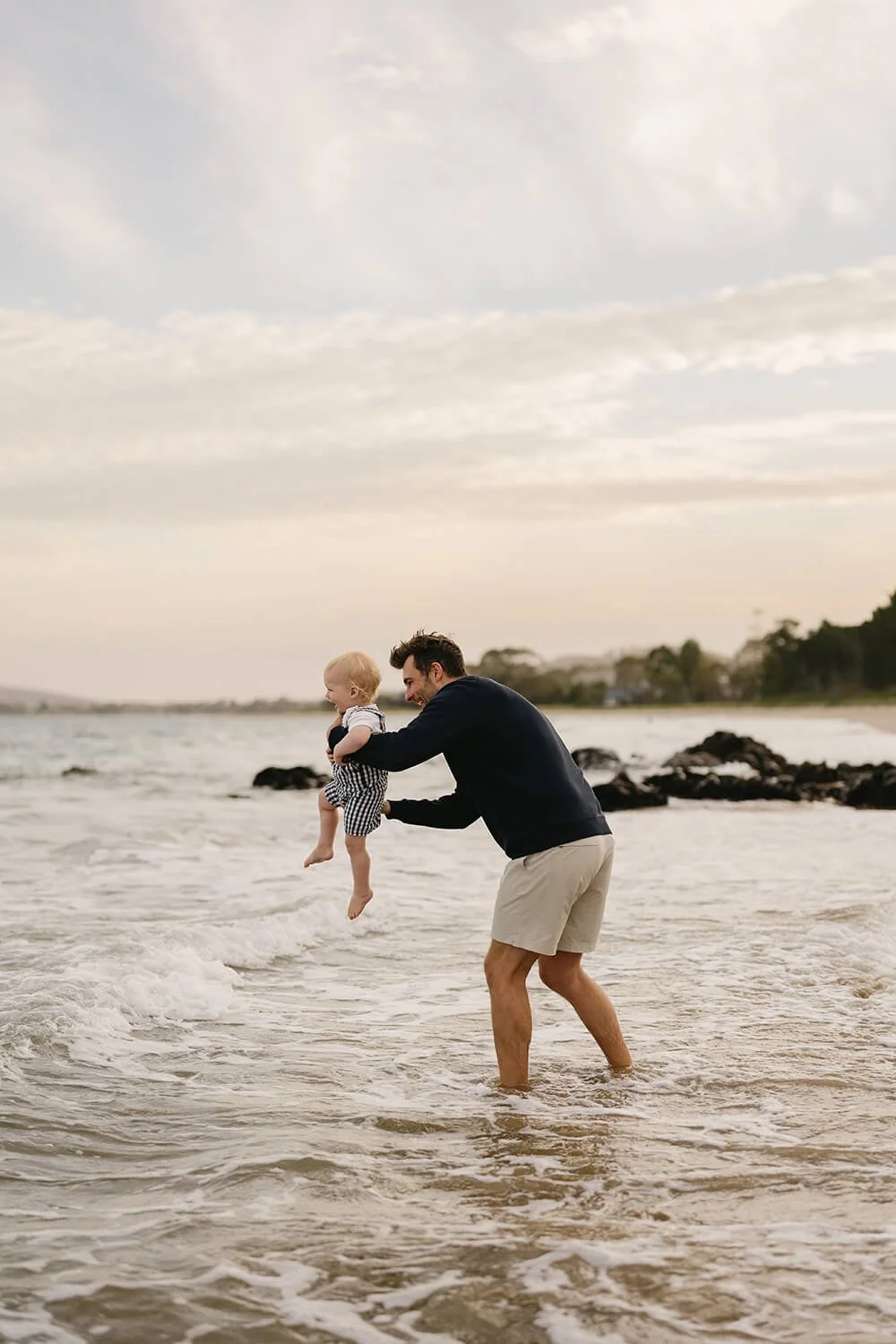 A man in shorts and a dark jacket playing with a young child in striped clothing at the beach, lifting the child over the water with a happy expression on both faces. The sky is partly cloudy, and rocky formations are visible in the background.