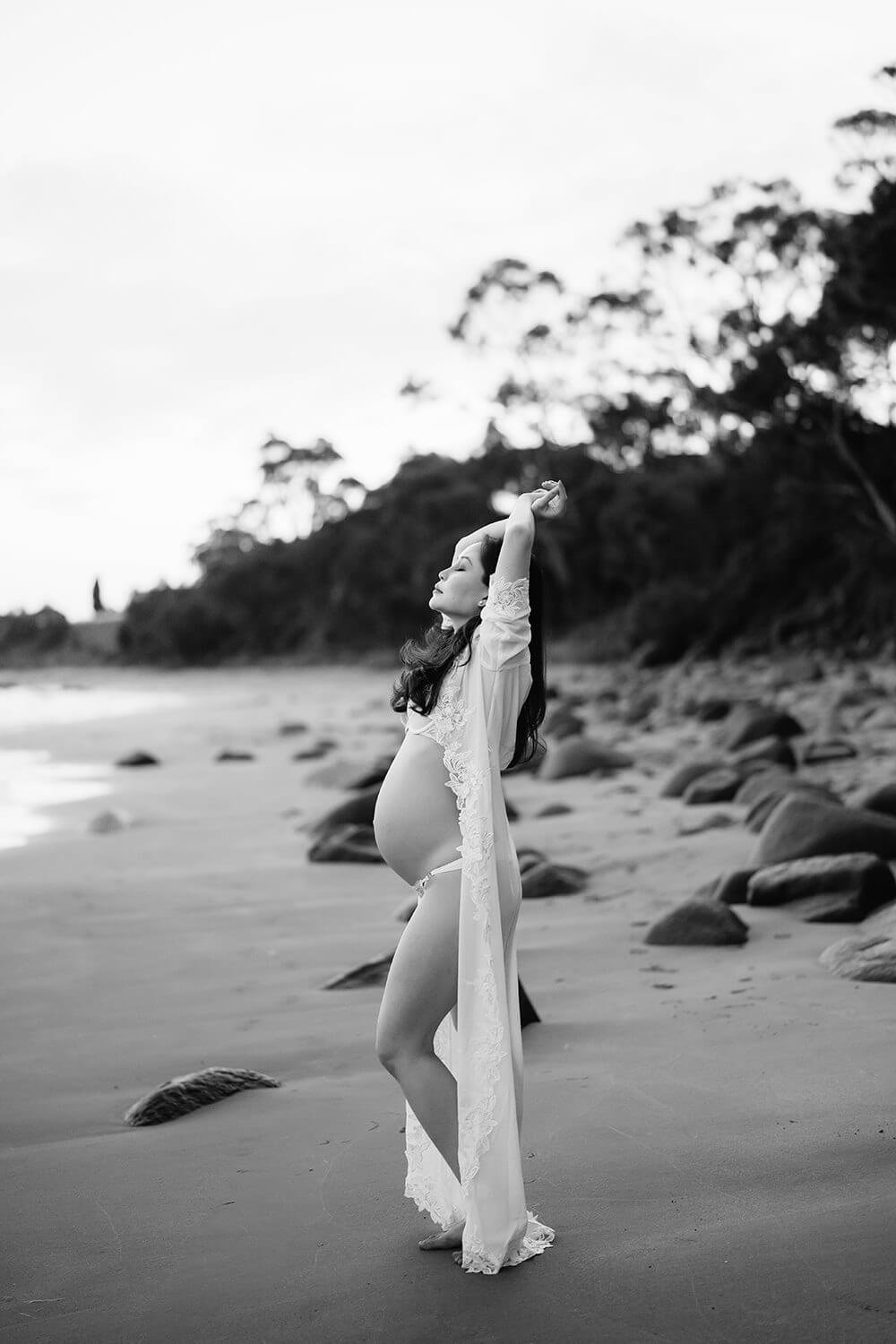 A pregnant woman in a lace robe standing barefoot on a beach, with her arms raised above her head, facing sideways with her eyes closed.