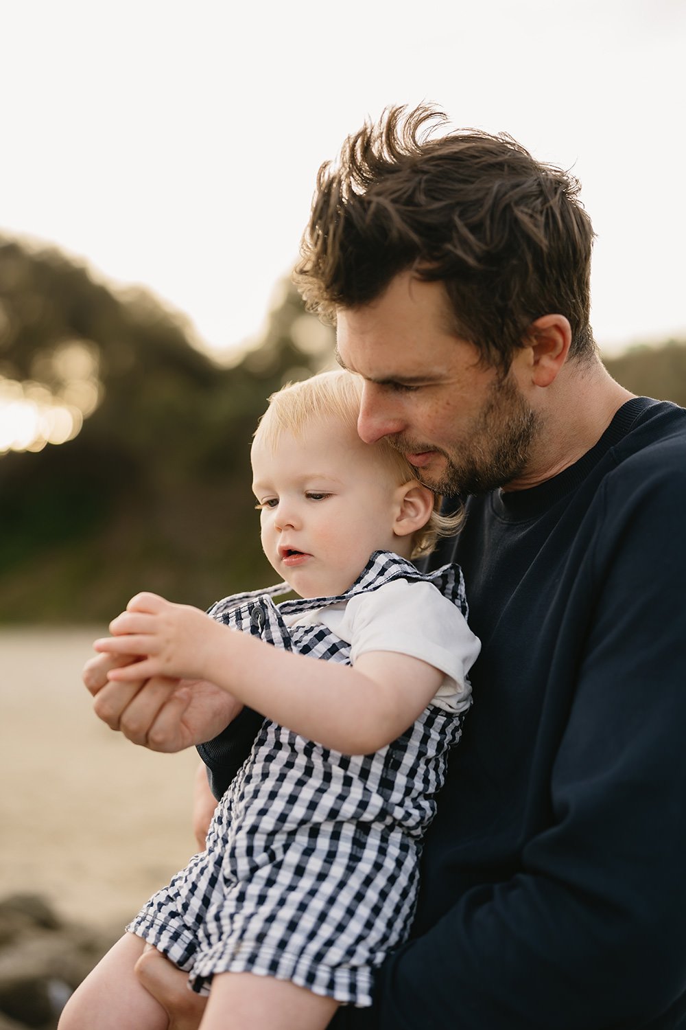 A man holding a young boy outdoors during sunset, both looking at something in the boy's hands.