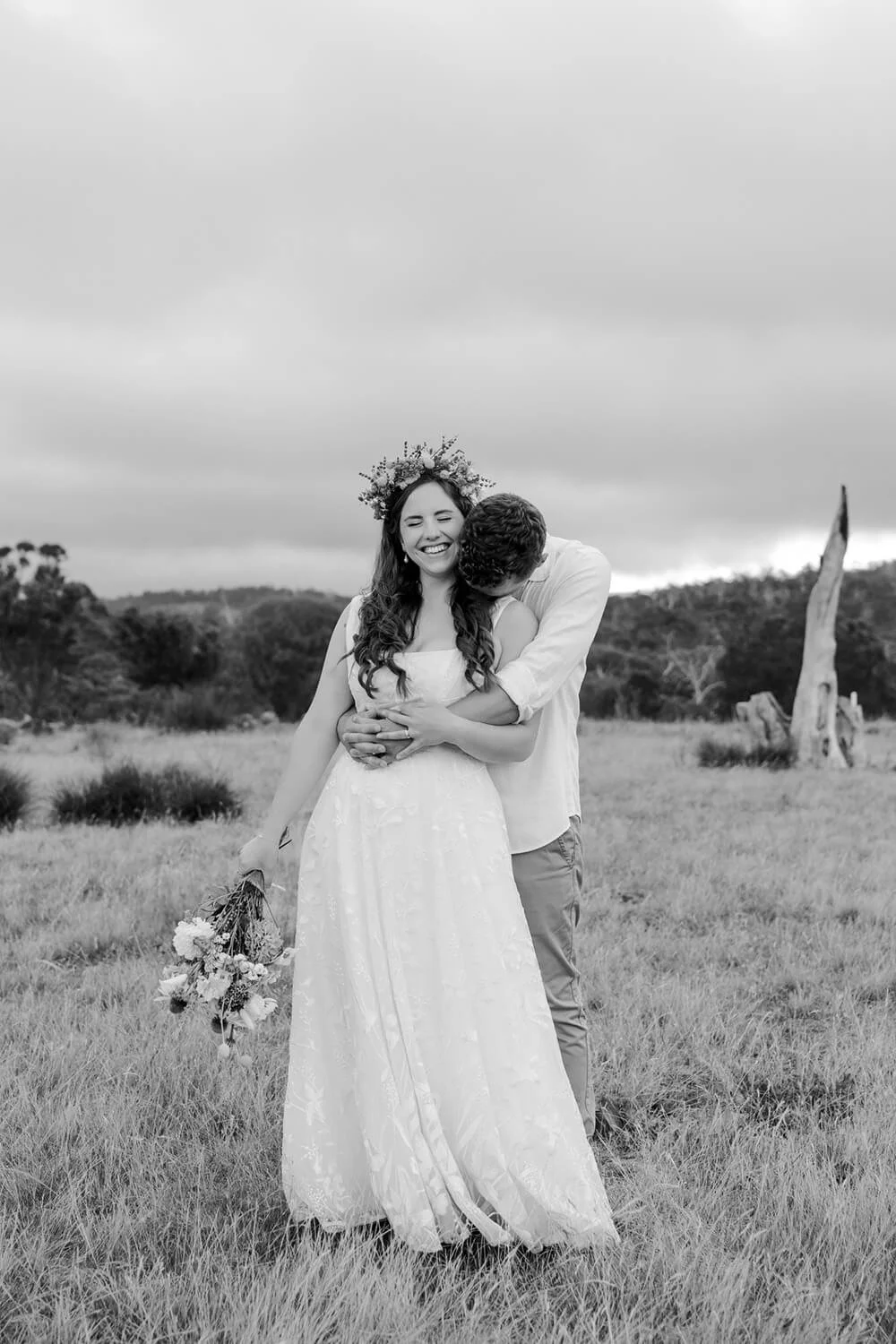 A black and white photo of a couple outdoors, with the woman wearing a wedding dress and floral crown, and the man hugging her from behind, both smiling and the woman holding a bouquet.