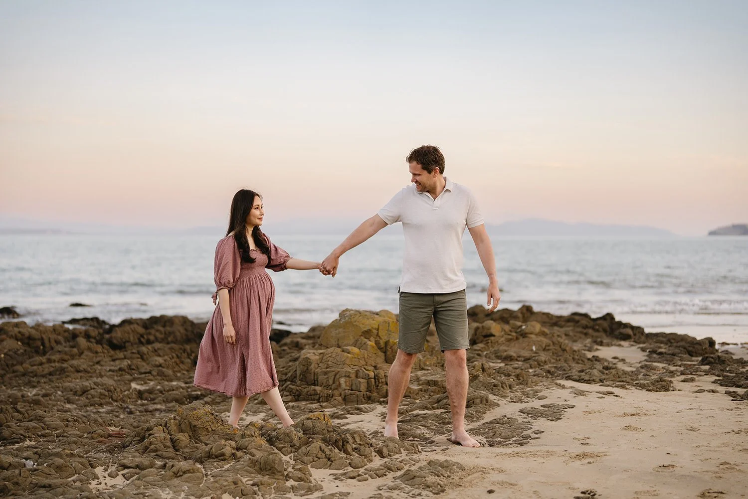 A couple holding hands on a beach with rocks and the ocean in the background, during sunset.