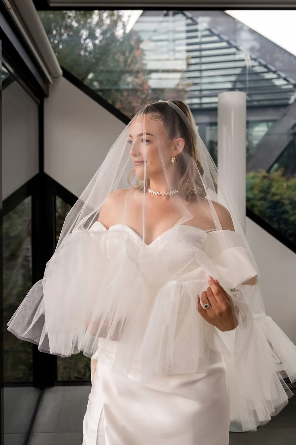 A bride in a white wedding gown with tulle details and a veil, standing inside a modern building with glass walls and a view of greenery outside.