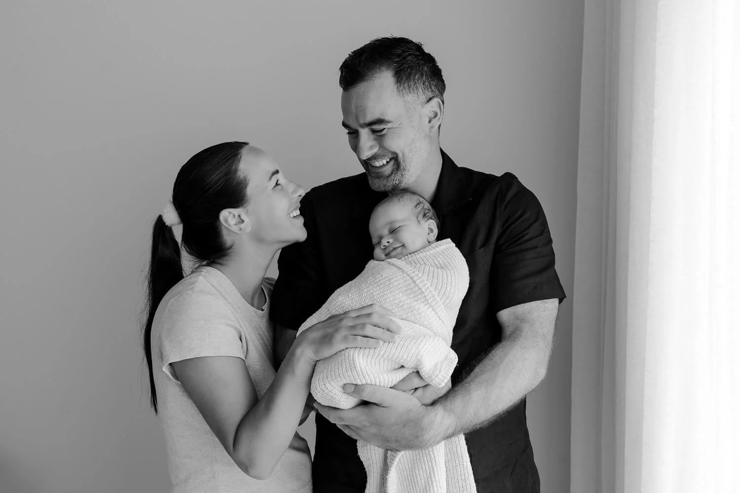 A happy family with a woman, a man, and a newborn baby. The woman and man are smiling at each other while holding the baby, who is wrapped in a blanket and appears to be sleeping or resting peacefully.