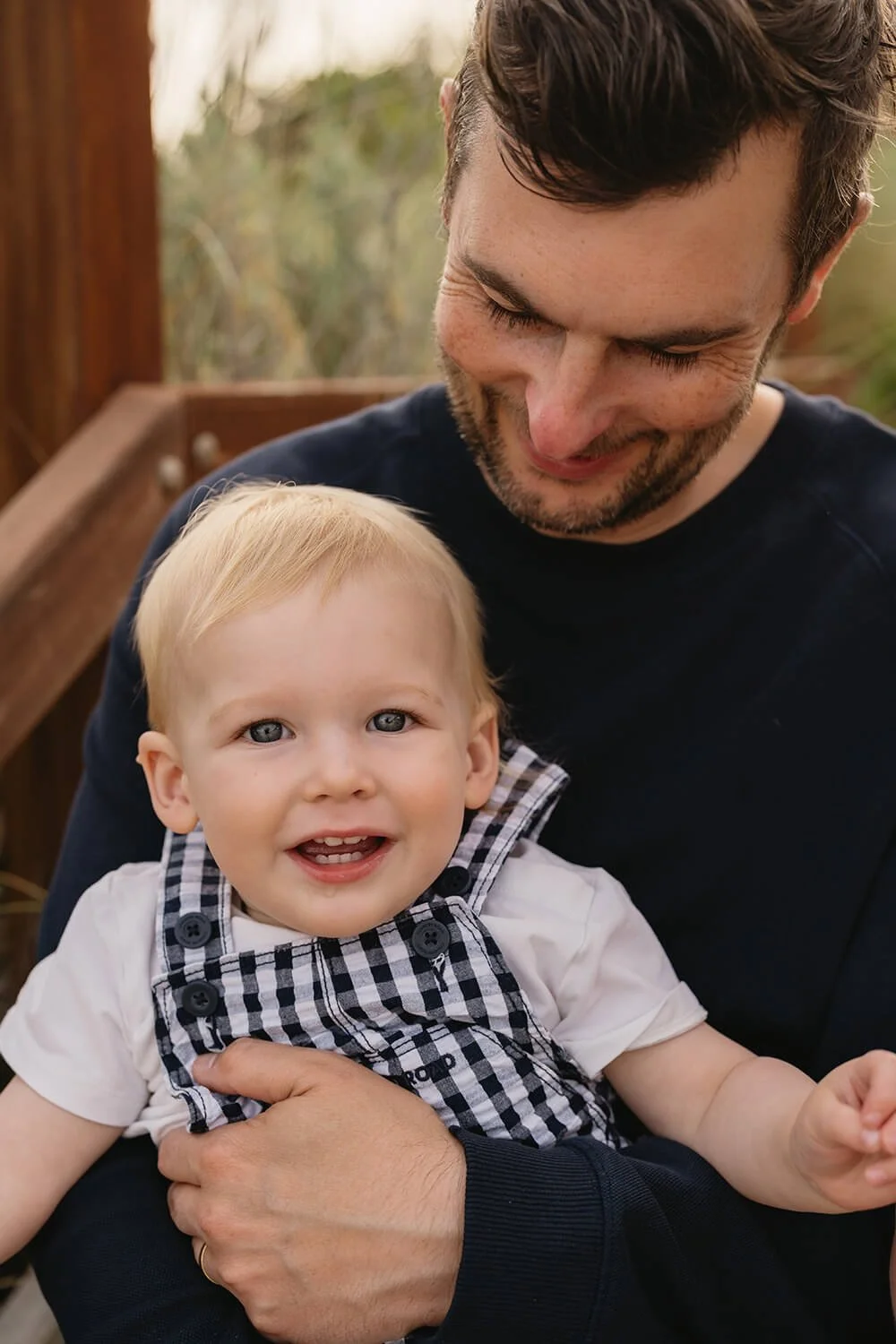 A smiling young man in a black shirt holding a toddler boy with blonde hair and blue eyes, wearing a white shirt and a black-and-white checkered vest, outside on a wooden deck with blurred trees in the background.