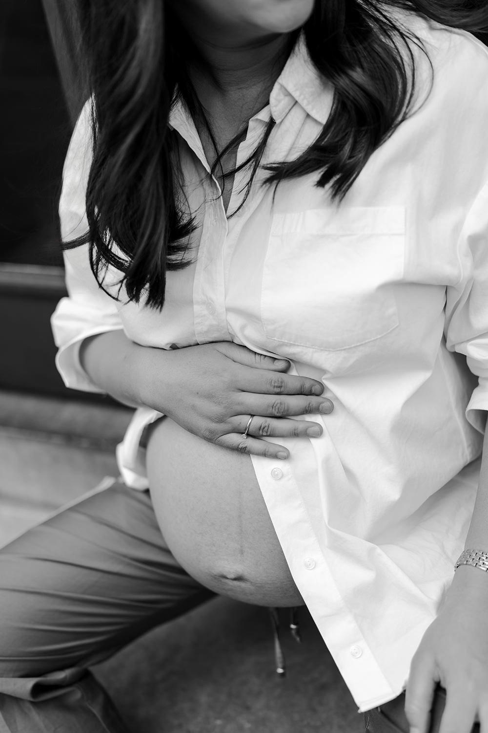 A woman with long dark hair, wearing a white shirt and pants, is sitting and holding her pregnant belly with both hands, in black and white.