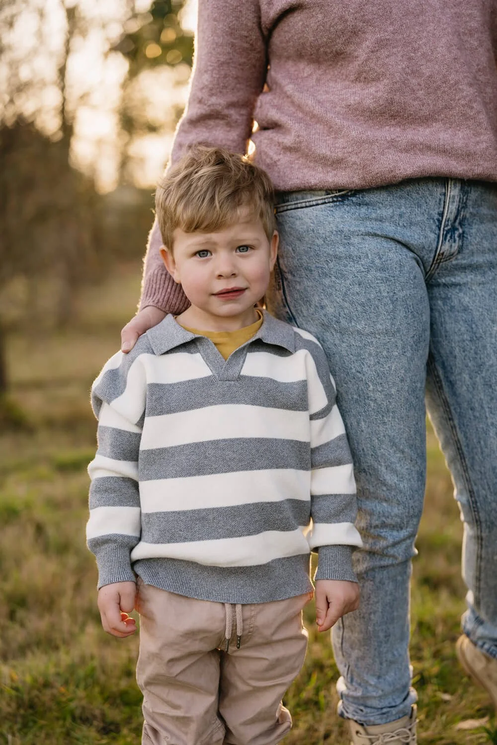 A young boy standing outdoors in front of a woman, possibly his mother, with both sunlight and trees in the background. The boy has short, light brown hair, wears a gray and white striped sweater, beige pants, and looks at the camera with a neutral e
