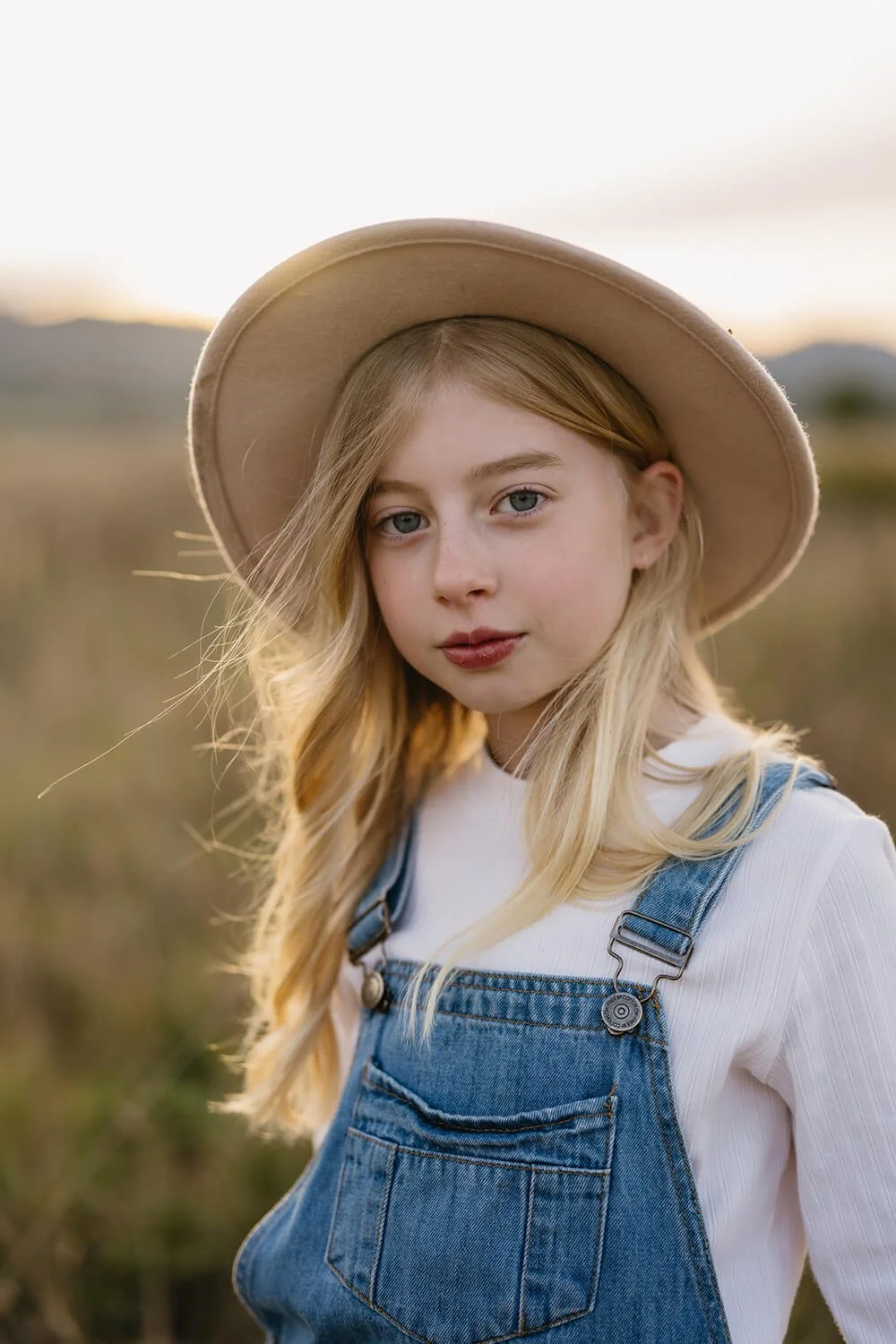 A young girl with long blonde hair, blue eyes, and fair skin wearing a beige wide-brimmed hat, white shirt, and denim overalls standing outdoors at sunset.