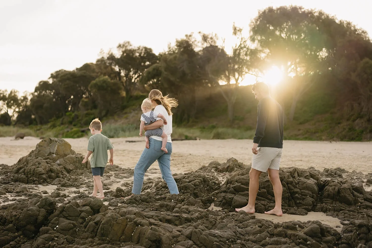 A family of four walking on a rocky beach at sunset, with trees in the background and the sun shining through the trees.