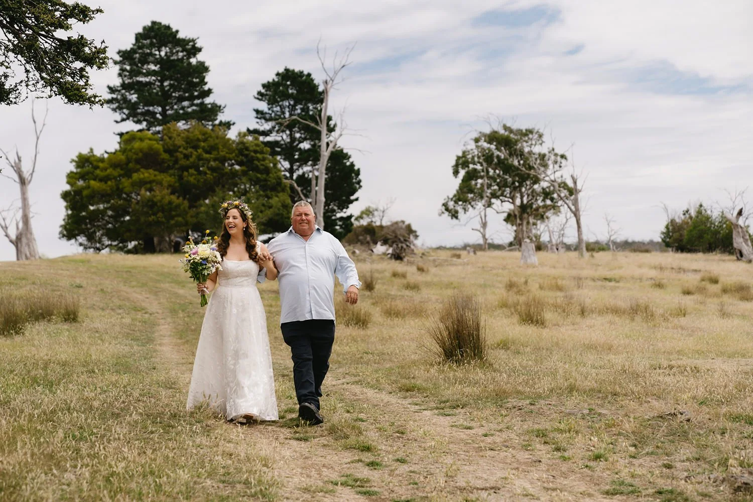 A bride in a white wedding dress with a floral crown holding a bouquet walking hand-in-hand with an older man in a white shirt in an open field with trees and cloudy sky.