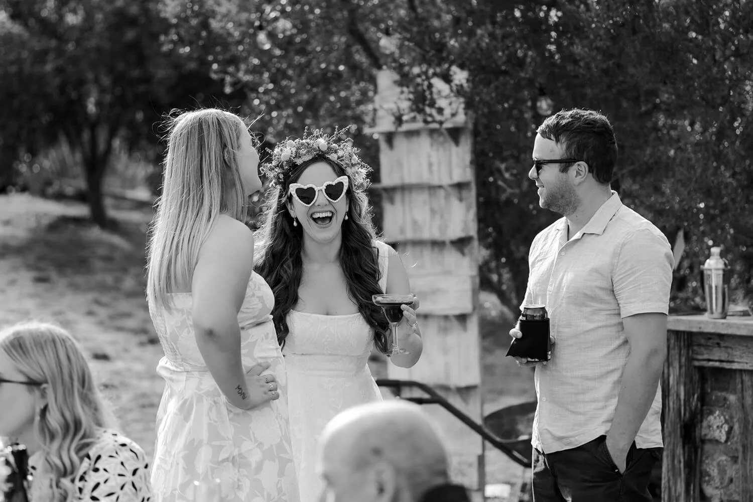 A black-and-white photo of three people outdoors, smiling and laughing. One woman is wearing heart-shaped sunglasses and a floral crown, holding a drink. Another woman is leaning in to talk to her, and a man is standing on the right, smiling and hold