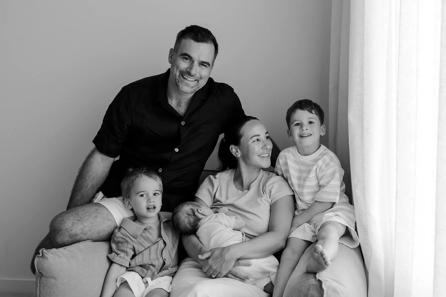 Black and white photo of a happy family sitting on a sofa near a window, including a man, a woman, three children, and a newborn.