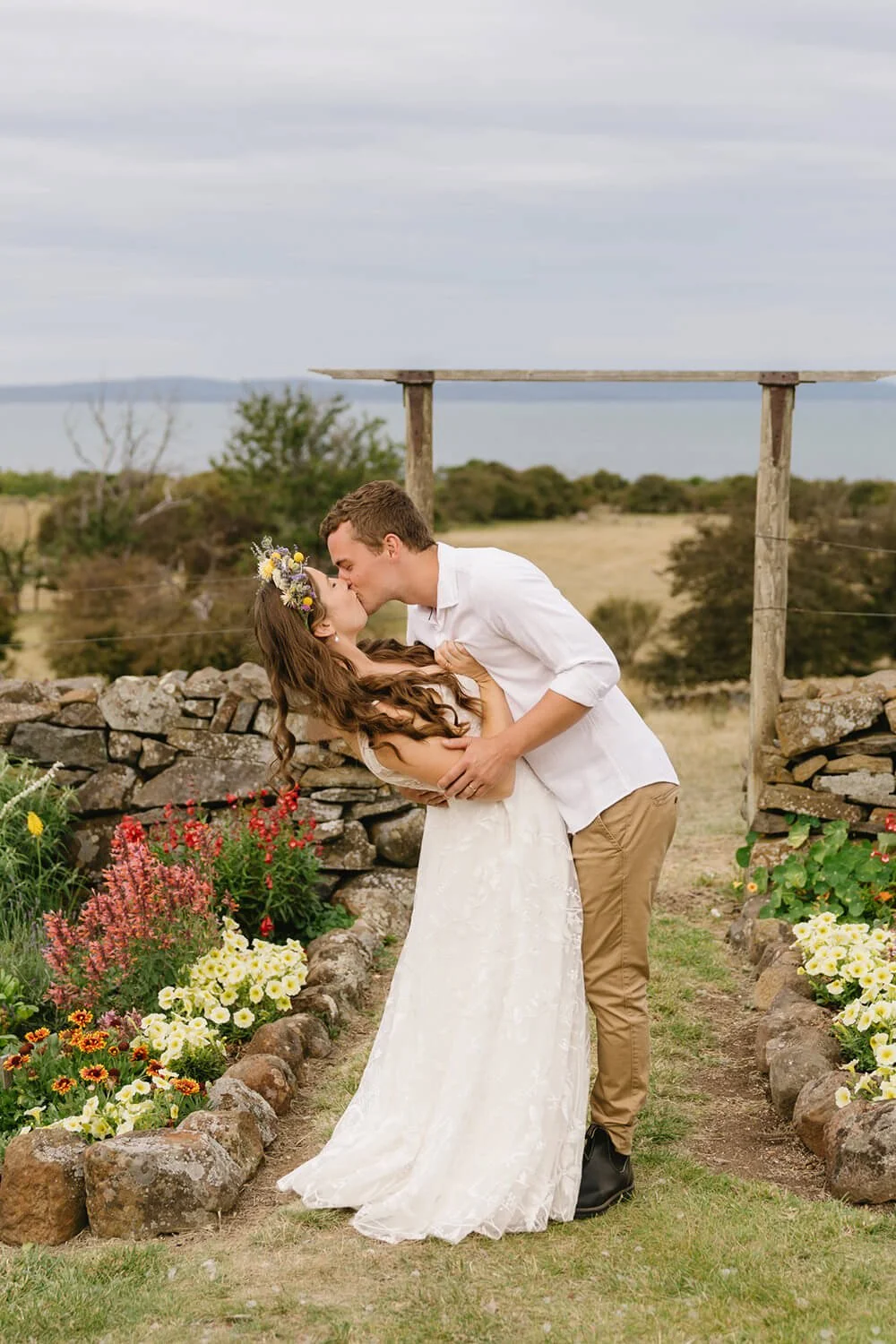 A couple sharing a kiss outdoors on a rocky garden path with flowers and a stone wall, overlooking water and a cloudy sky.