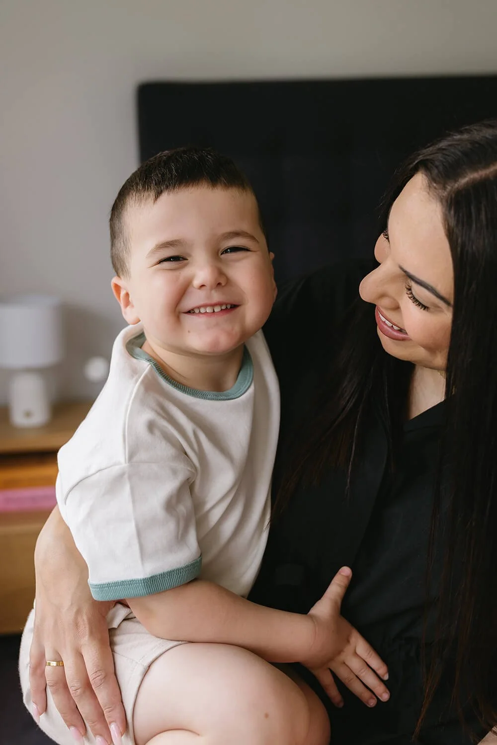 A smiling young boy with short dark hair sitting on a woman's lap, looking at the camera. The woman, with long dark hair, is smiling and looking at the boy. They are indoors, with a blurred background.