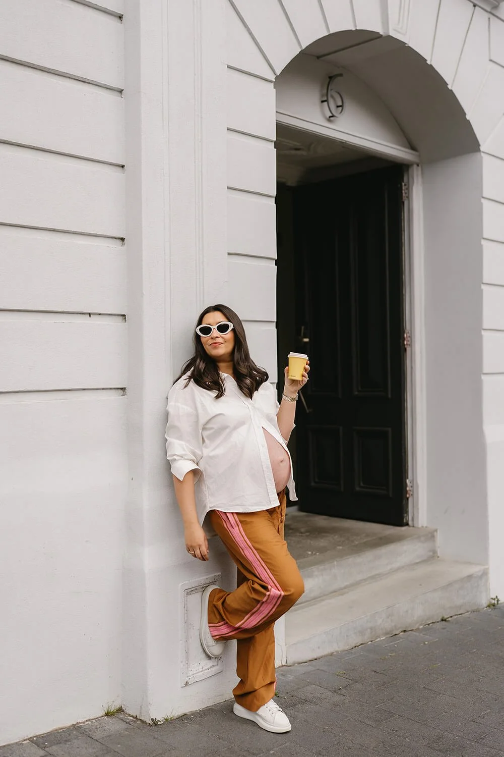 A pregnant woman stands against a white building wall, holding a coffee cup, wearing white sunglasses, a white unbuttoned shirt, brown pants with pink stripes, and white sneakers.