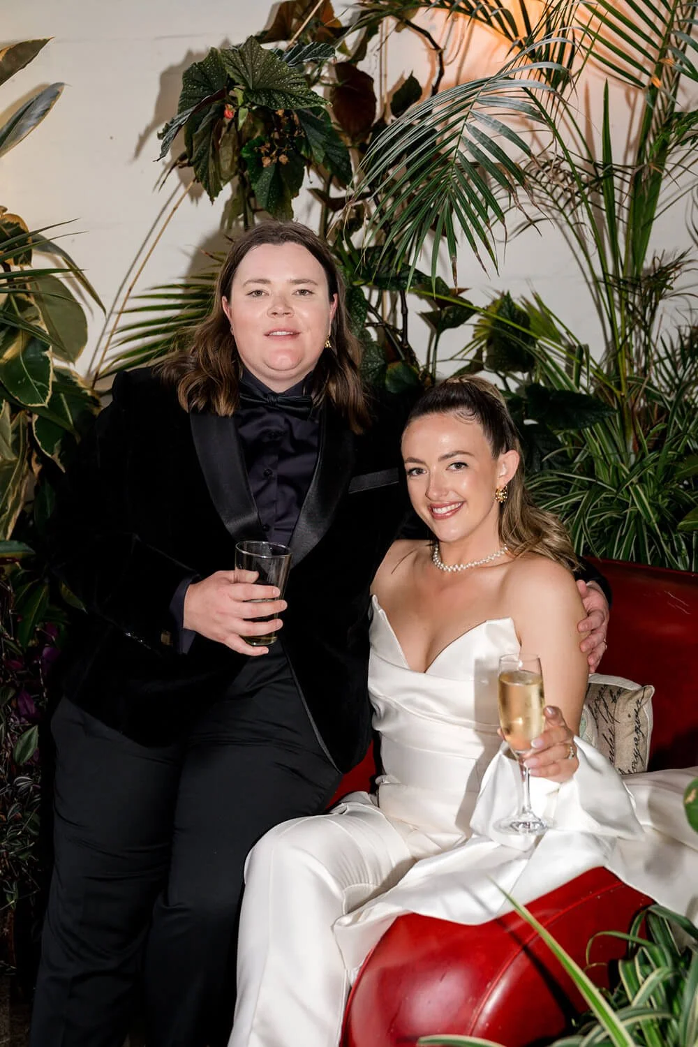 A man and a woman posing together at a formal event, surrounded by green plants. The woman is wearing a white strapless gown and holding a glass of champagne, while the man is dressed in a black tuxedo with a bow tie and holding a drink.