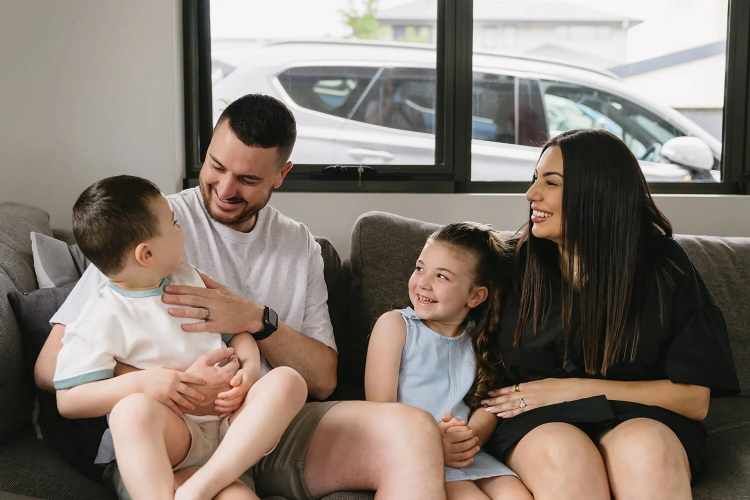 A happy family of four sitting on a couch in a living room, smiling and engaging with each other. The father is holding a young boy, and the mother and daughter are sitting nearby. Behind them is a window revealing parked cars outside.