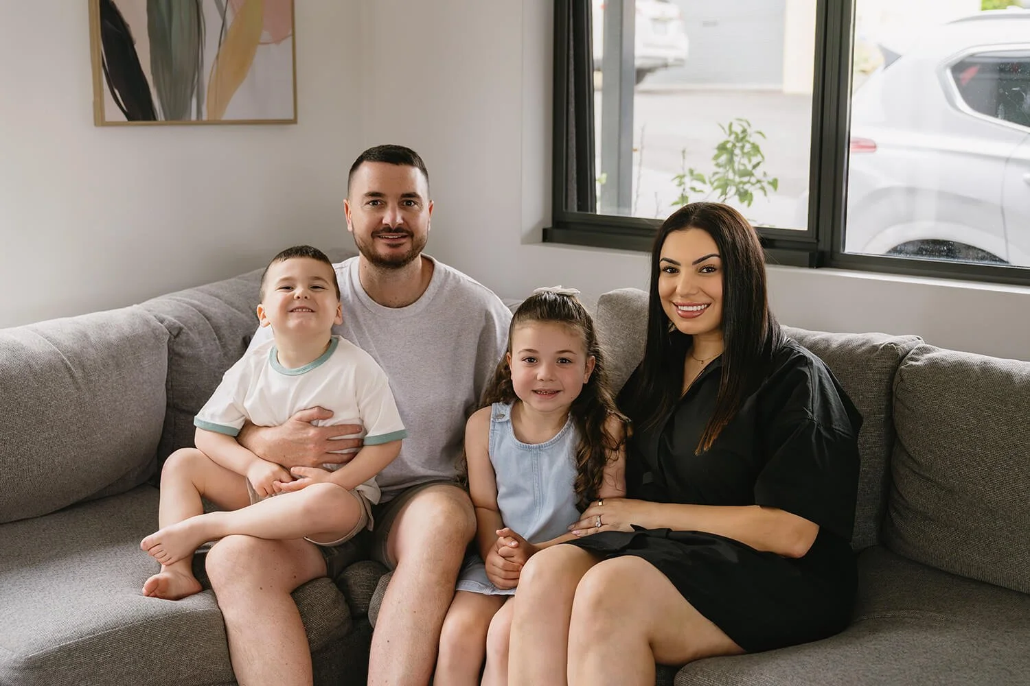 A family of four sitting on a gray couch in a living room, smiling at the camera. The father has short dark hair and a beard, wearing a light gray T-shirt. The mother has long dark hair, wearing a black dress. The young boy has short hair, wearing a 