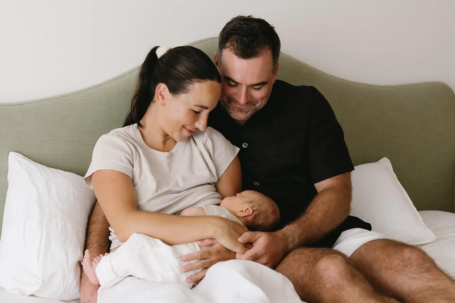 A family of three, a woman, a man, and a newborn baby, sitting together on a bed, with the woman breastfeeding the baby and everyone smiling