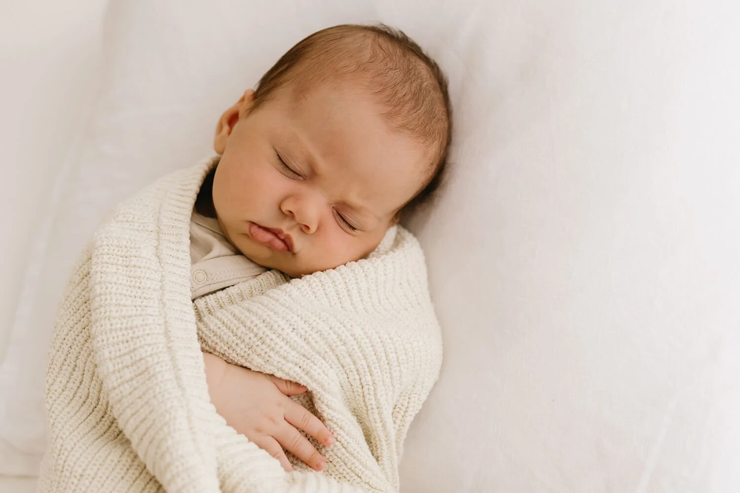 A sleeping baby with light skin and brown hair, wrapped in a cream-colored knit blanket, resting on a white pillow and bed.