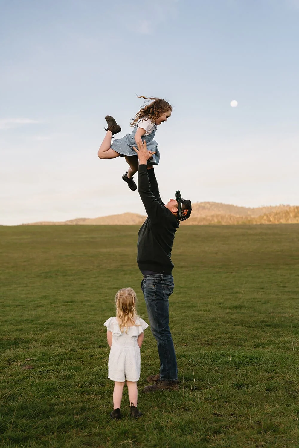 A man wearing a cap lifts a young girl into the air in a grassy field, with a second young girl watching nearby. The sky is clear with the moon visible, and rolling hills are in the distance.