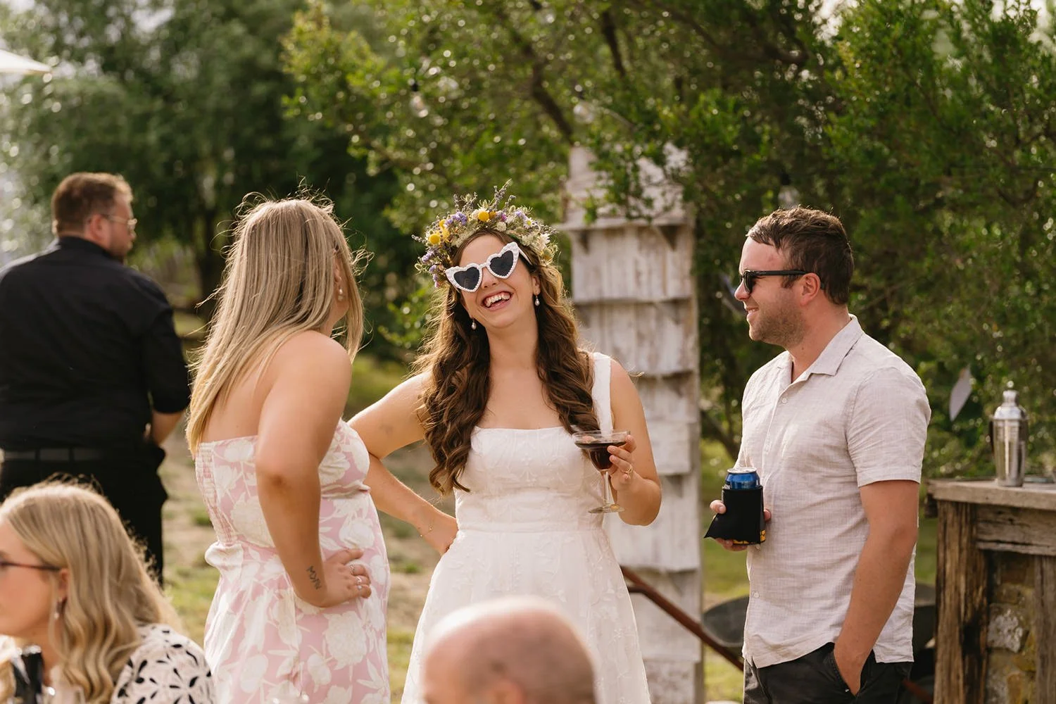 Group of people enjoying an outdoor celebration or wedding, with a woman wearing a flower crown and heart-shaped sunglasses, holding a wine glass, and smiling while talking to a man and another woman in a garden setting.