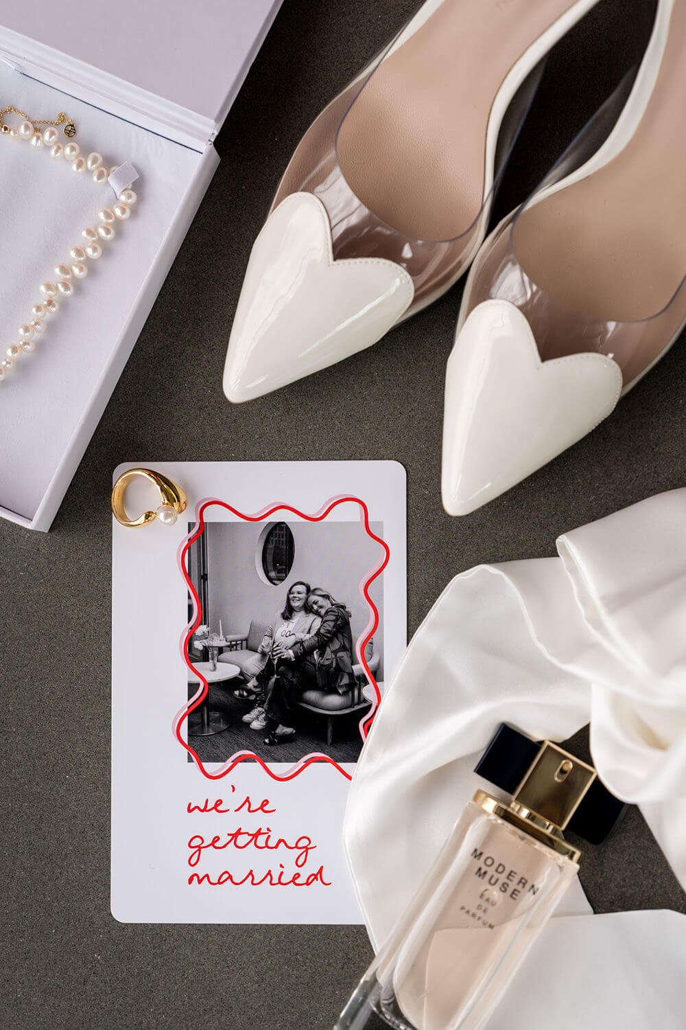 Wedding invitation, a pearl necklace, a gold ring, a perfume bottle, a pair of beige and clear high heel shoes with white hearts, and a black-and-white photo with red text that says 'we're getting married' on a dark surface.