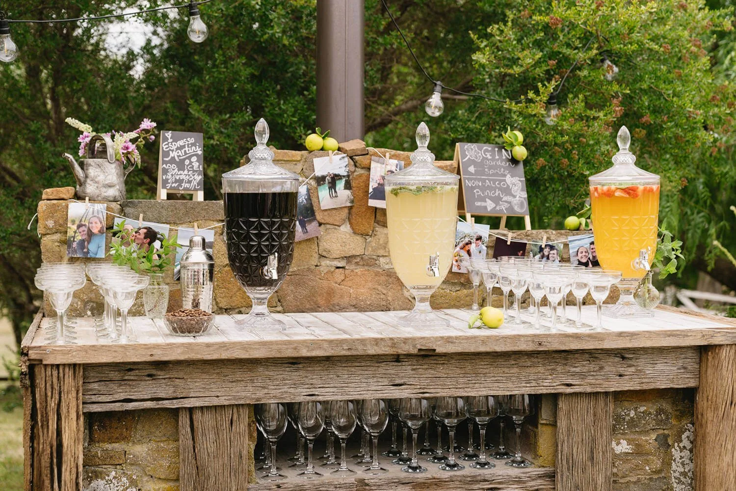 A rustic outdoor drink station with three large glass dispensers filled with dark, yellow, and orange beverages, surrounded by small chalkboard signs, photo strings, and a stone wall backdrop with greenery and string lights.