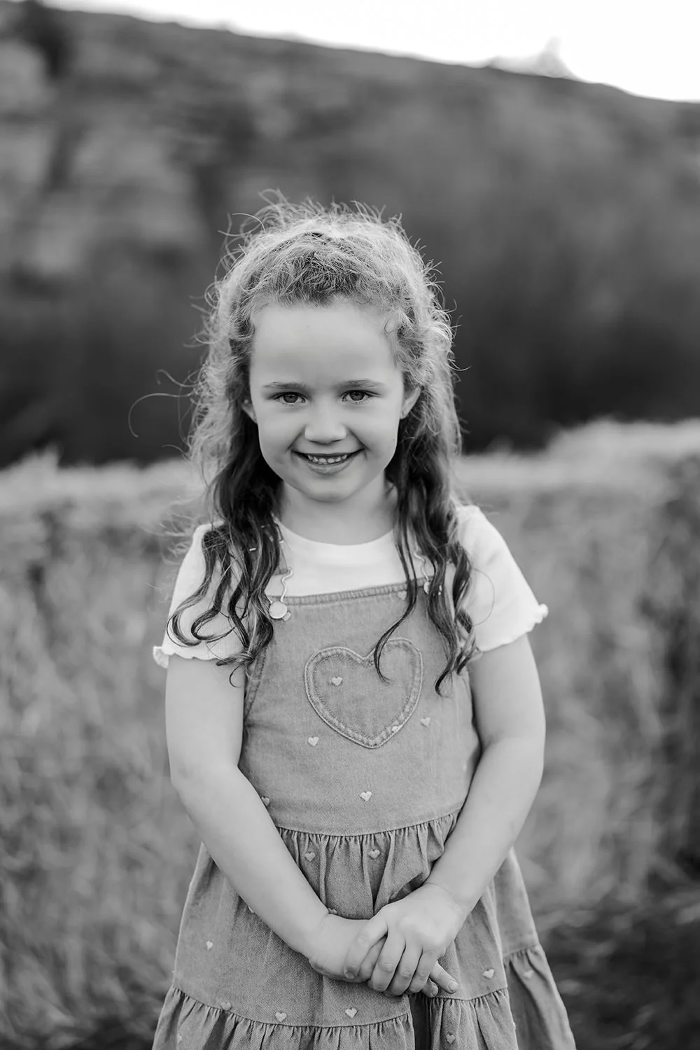 A young girl with curly hair smiling outdoors, wearing a dress with a heart embroidery.