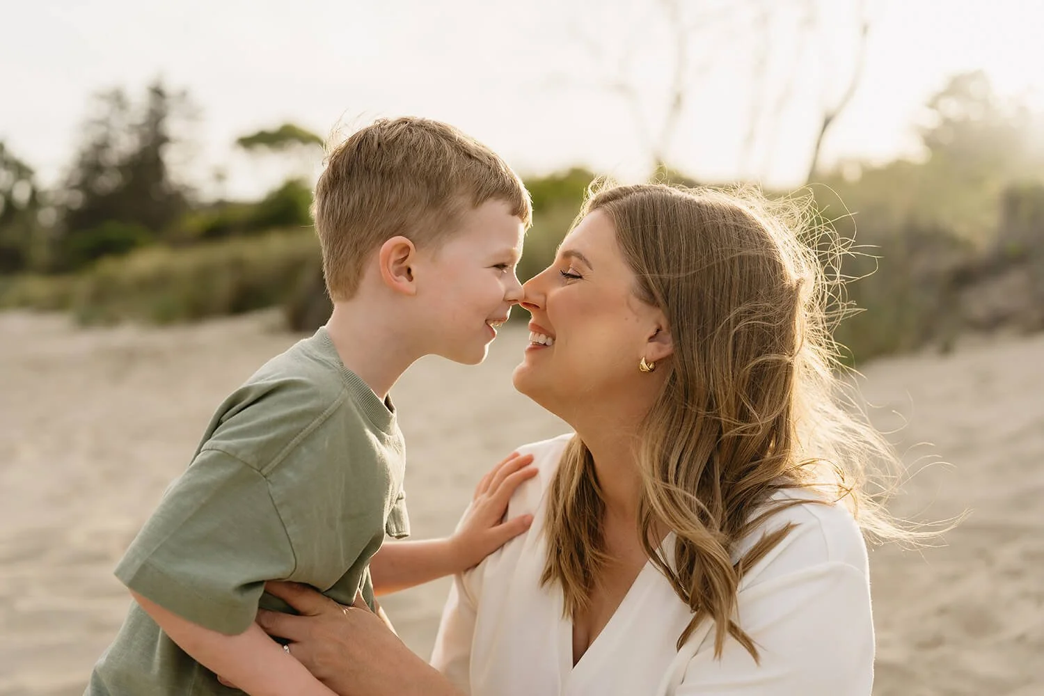 A woman and a boy smiling nose-to-nose on a beach during sunset, with trees in the background.