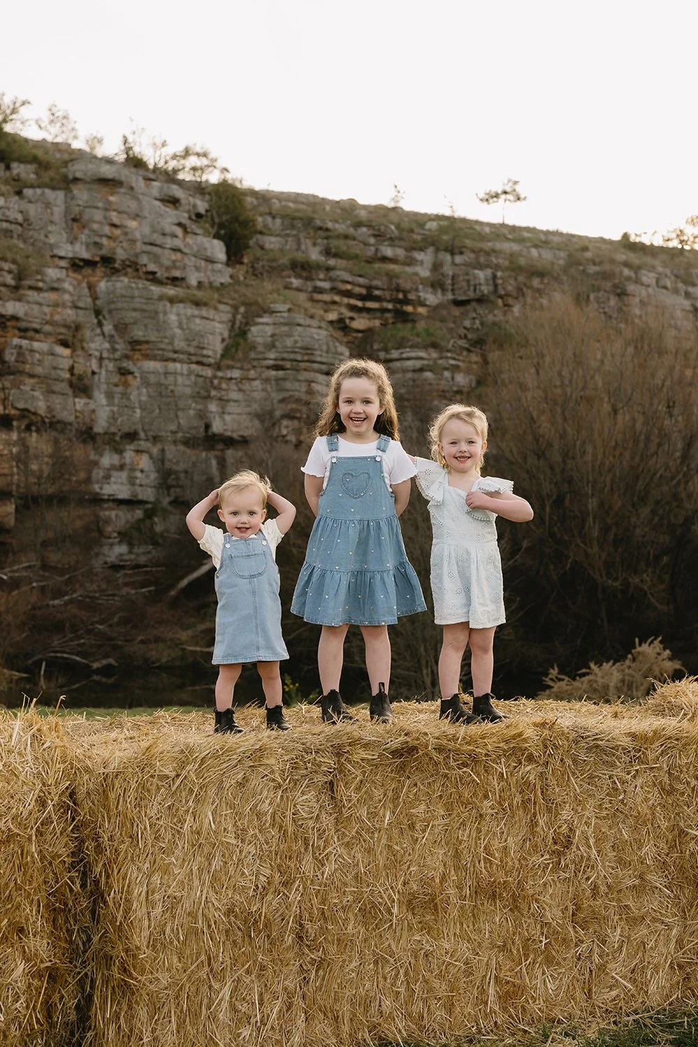 Three young girls wearing dresses standing on a haystack outdoors near rocky cliffs, smiling and posing for the camera.