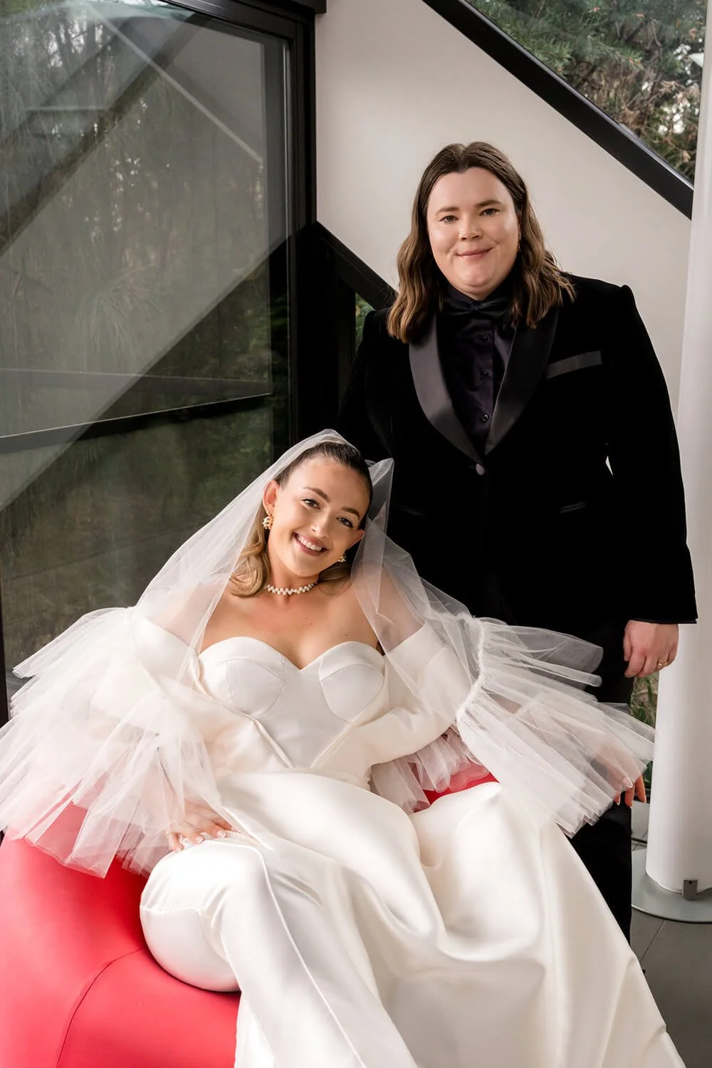 A woman sitting in a wedding dress and veil, smiling, with a person in a black tuxedo standing next to her, indoors near a window.