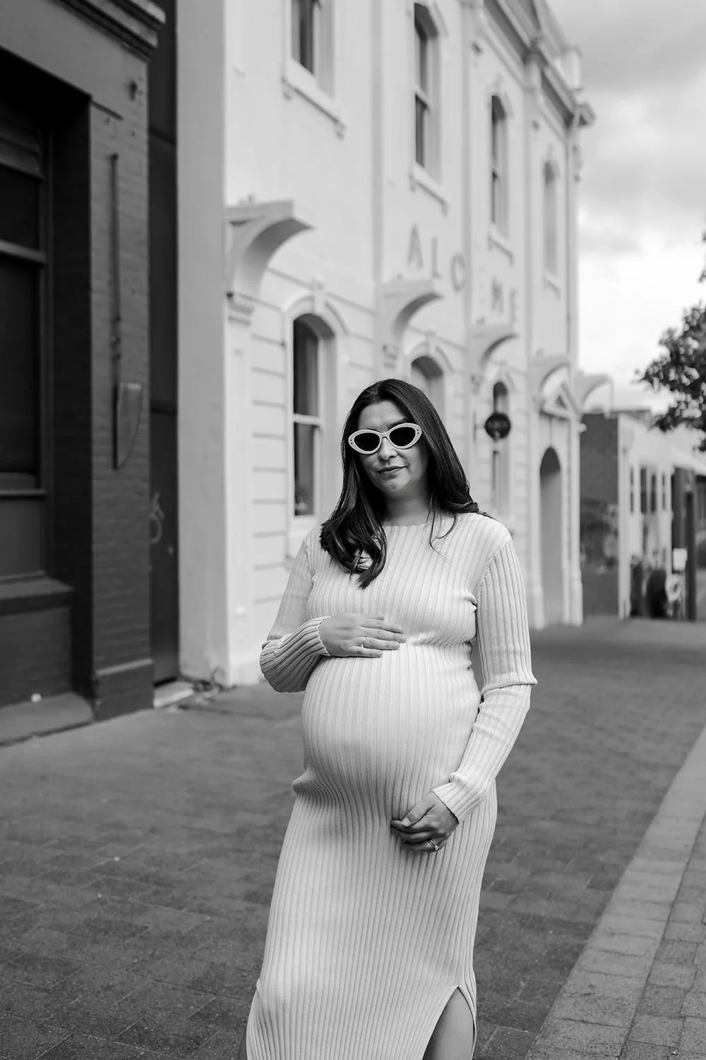 A pregnant woman wearing sunglasses and a ribbed dress standing on a city sidewalk with buildings and trees in the background.