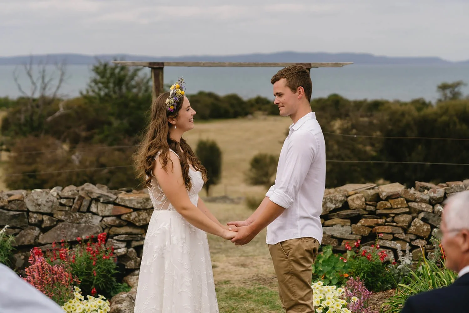 A woman in a white dress and floral crown holding hands with a man in a white shirt and khakis during a wedding ceremony outdoors near a stone wall, with flowers and landscape in the background.